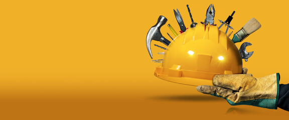 Manual worker with protective work gloves holding a yellow safety helmet with many work tools, on a yellow and orange background with copy space and reflections. - obrazy, fototapety, plakaty