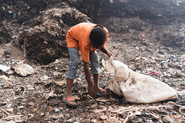 Young garbage picker working hard in an African landfill amidst dirt and toxic materials putting...