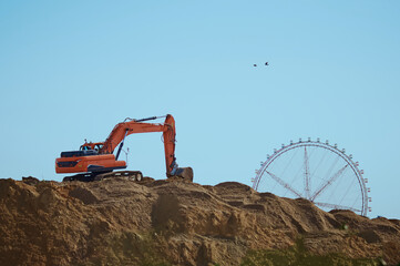 An orange excavator is excavating on a hill against the backdrop of a ferris wheel and blue sky....