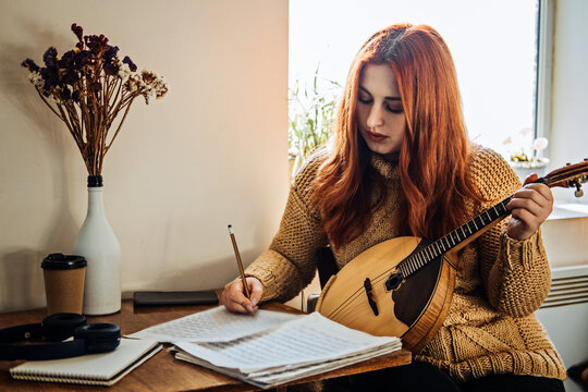 Redhead woman playing acoustic Domra Folk Musical Instrument and looking into note. Young woman taking music lessons at home
