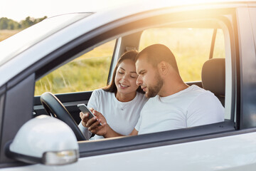 Portrait of man and woman in a new car, traveling together, wife holding smart phone and showing...