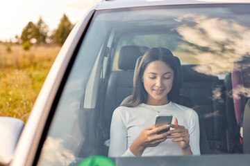 Smiling beautiful young adult woman looking at her mobile phone while sitting inside car, attractive dark haired female wearing white shirt in her automobile surfing internet via smart phone.