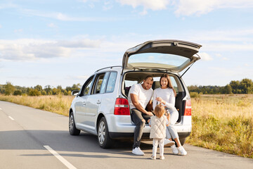 Outdoor shot of happy couple with their infant daughter sitting in car trunk and drinking hot beverage, coffee or tea from thermos, enjoying to have journey together.
