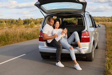 Horizontal shot of romantic young couple sitting together in the car trunk with cell phone on the roadside, using smart phone while traveling by car, enjoying their journey.