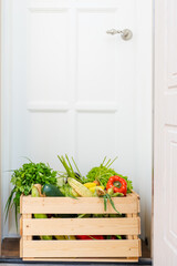 Wooden box full of fresh vegetables, fruits and greens stand on doorstep of house near white door....