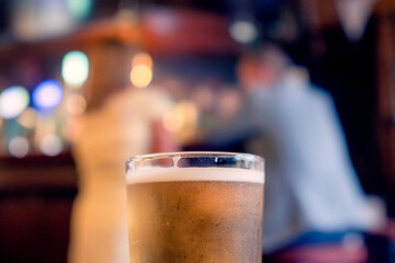 Pint glass of cold lager beer of cider in focus. Well dressed woman and man out of focus in the background. Date of flirt concept. Enjoy cold craft alcohol drink in a traditional Irish pub.