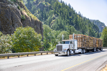 Classic old style white big rig bonnet semi truck tractor transporting wood pallets on two flat bed...