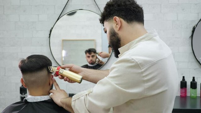 A male barber cuts a client's hair in a modern barbershop using an electric razor, he sits in a chair in front of a mirror.