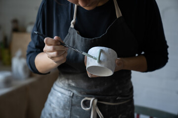 Woman enjoys new creative hobby painting white ceramic mug with paintbrush standing on blurred background. Female person in black apron takes classes of handmade pottery craft in workshop closeup