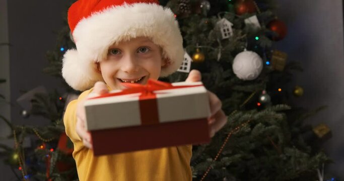 Child holds out and gives you gift. Video portrait of boy in Santa Claus hat with gift box in hands