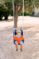 Little serious girl in sunglasses sits on a swing holding on the ropes. High quality photo