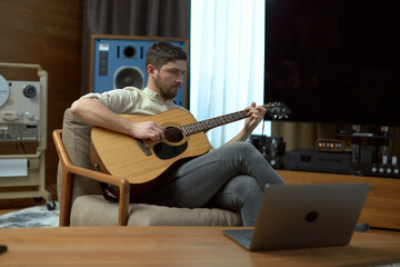 Musician playing acoustic guitar music at home, using laptop computer for online musical concert or distant education