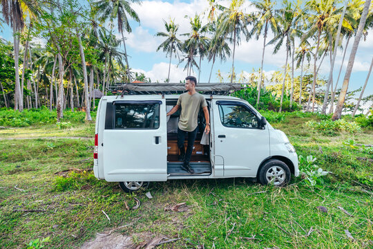 Young Man Exiting White Camper Van Parked In A Grassy Green Field Surrounded By Tropical Trees In Bali Indonesia On Sunny Day