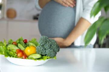 Pregnant woman with bowl of fresh vegetables, broccoli, leaf salad, tomato, cucumber on table....