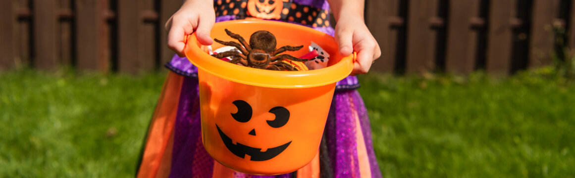 cropped view of girl holding  trick or treat bucket with sweets and toy spider, banner