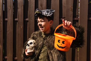 excited asian boy in werewolf costume holding halloween bucket and spooky skull while looking away