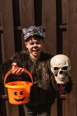 happy asian boy in werewolf costume holding spooky skull and halloween bucket while looking at...