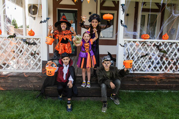 multiethnic kids in halloween costumes grimacing and showing scary gestures near decorated cottage