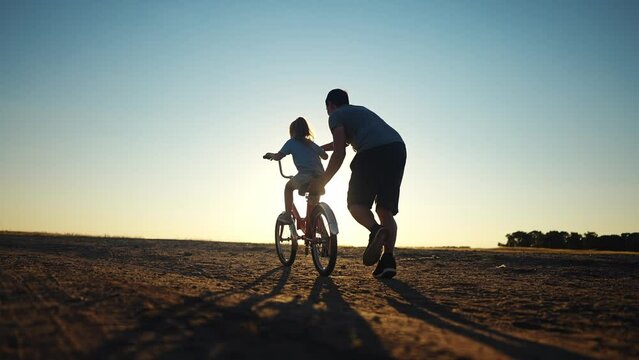 dad teaches daughter to ride a bike. happy family childhood dream concept. father and little daughter learn to ride a bike silhouette in the park. happy family goes in sunlight for sports outdoors