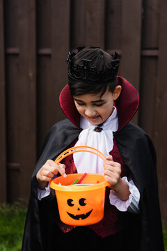 Asian Kid In Vampire King Costume Looking Into Trick Or Treat Bucket With Candies