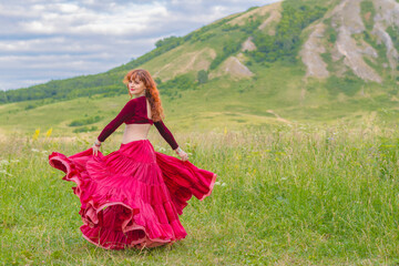 Young red-haired girl in red ball gown is dancing incendiary dance on green meadow against backdrop of relic mountain Shihan.