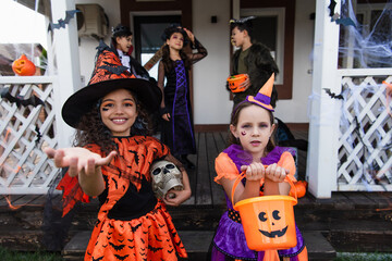 smiling girl in witch costume standing with outstretched hand near friend with halloween bucket