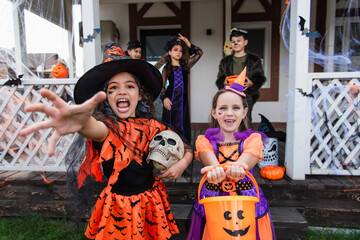 girl in witch costume screaming with outstretched hand near smiling friend with trick or treat bucket