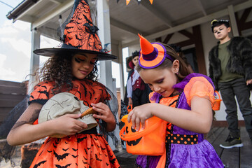 girl in witch costume feeding skull with cookie near friend looking into halloween bucket