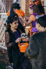 Cheerful kid in halloween costume holding skull and bucket with candies near asian friend outdoors