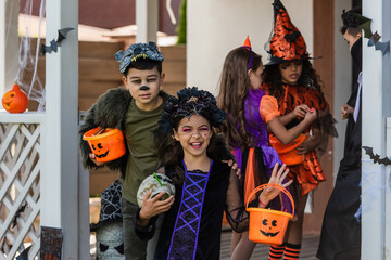 Happy girl in halloween costume holding skull near asian friend with bucket and kids in backyard