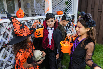 Obraz na płótnie Canvas Asian boy in costume looking at camera near cheerful friends with bucket talking during halloween celebration outdoors