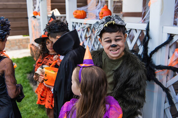 Asian kid in halloween costume grimacing at camera near friends and decor outdoors