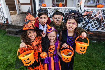 Multiethnic kids in halloween costumes holding buckets with candies and looking at camera in...