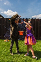 Kid in halloween costume standing near asian friend with bucket in backyard