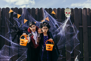 Interracial children in costumes holding halloween buckets near spider web on fence outdoors