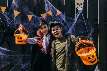Asian kids holding halloween buckets with candies near spider web on fence during party in backyard