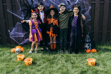 Smiling multiethnic kids in costumes looking at camera near halloween decor on fence in backyard