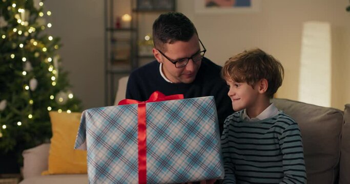 Curious child enjoys the surprise of a Christmas gift, unties the bow shakes the box with interest, dad looks at his son with joy and pride.