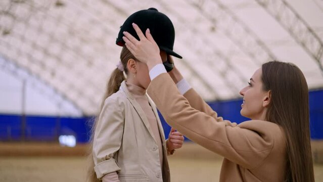 Long-haired woman in a beige coat kneeled near a young girl. Lady puts jockey hat on daughter's head but it's little too big.