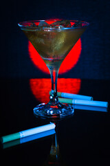 Cocktail glass with ice filled with alcohol. Cigarettes scattered around a cocktail glass with an iced drink. There's a glass and cigarettes on the bar.