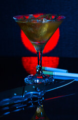 Art photo - still life with a glass, a knife and scattered cigarettes. A cocktail glass with alcohol on ice, a knife and two cigarettes on a dark background.