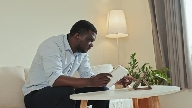 Medium shot of confident African American man in light blue office shirt taking notes in notebook while working on digital tablet sitting at desk in home office