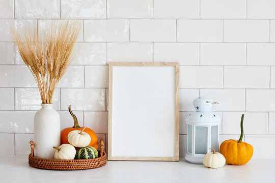 Autumn still life. Picture frame mockup, tray with vase of dry wheat, decorative pumpkins, lamp on white table in scandinavian kitchen interior. Fall, Thanksgiving holiday concept.