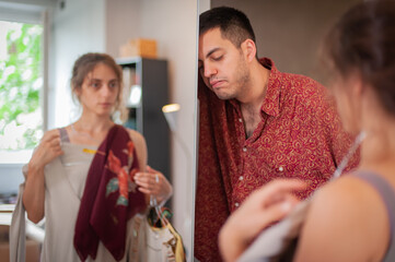 Charming young woman is trying on clothes and having fun standing in front of the mirror in the bedroom, but the man is exhausted and bored