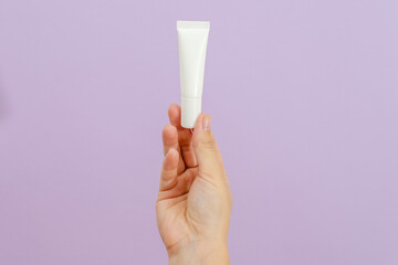 Hand holds plastic white tube isolated on lilac background. Beauty concept. Packaging tube for cosmetic products