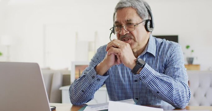 Video of happy senior biracial man sitting at desk using headset and laptop making video call