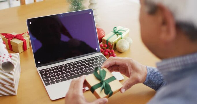 Video of senior biracial man holding gift making christmas laptop video call, copy space on screen