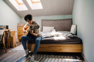 Young asian man in headphones playing guitar while sitting on bed