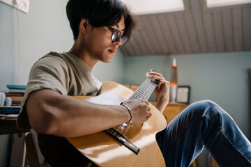Young asian man in headphones playing guitar while sitting on chair