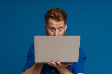 Ginger shocked man looking at camera while posing with laptop